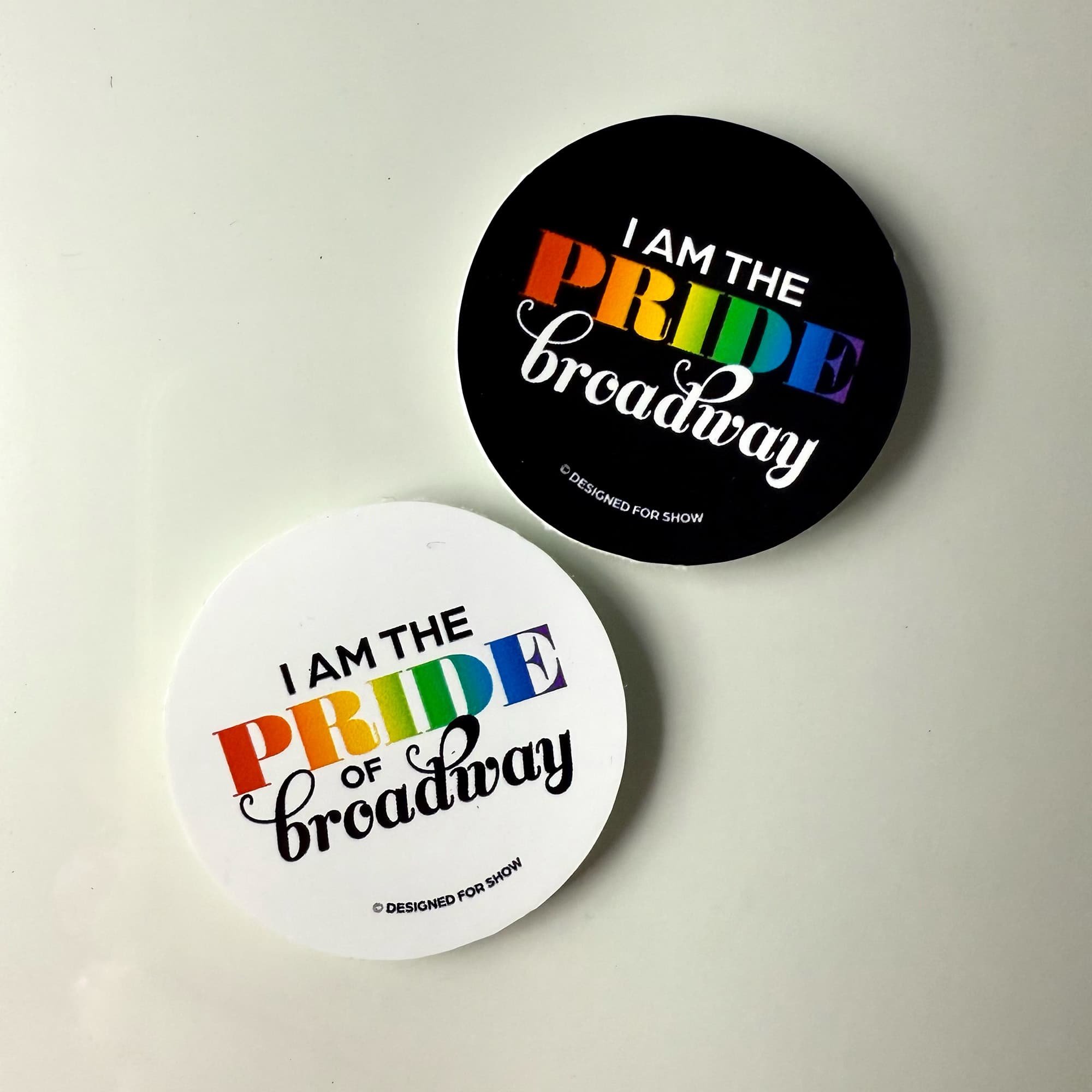 Two stickers, one black, one white, that say I am the Pride of Broadway