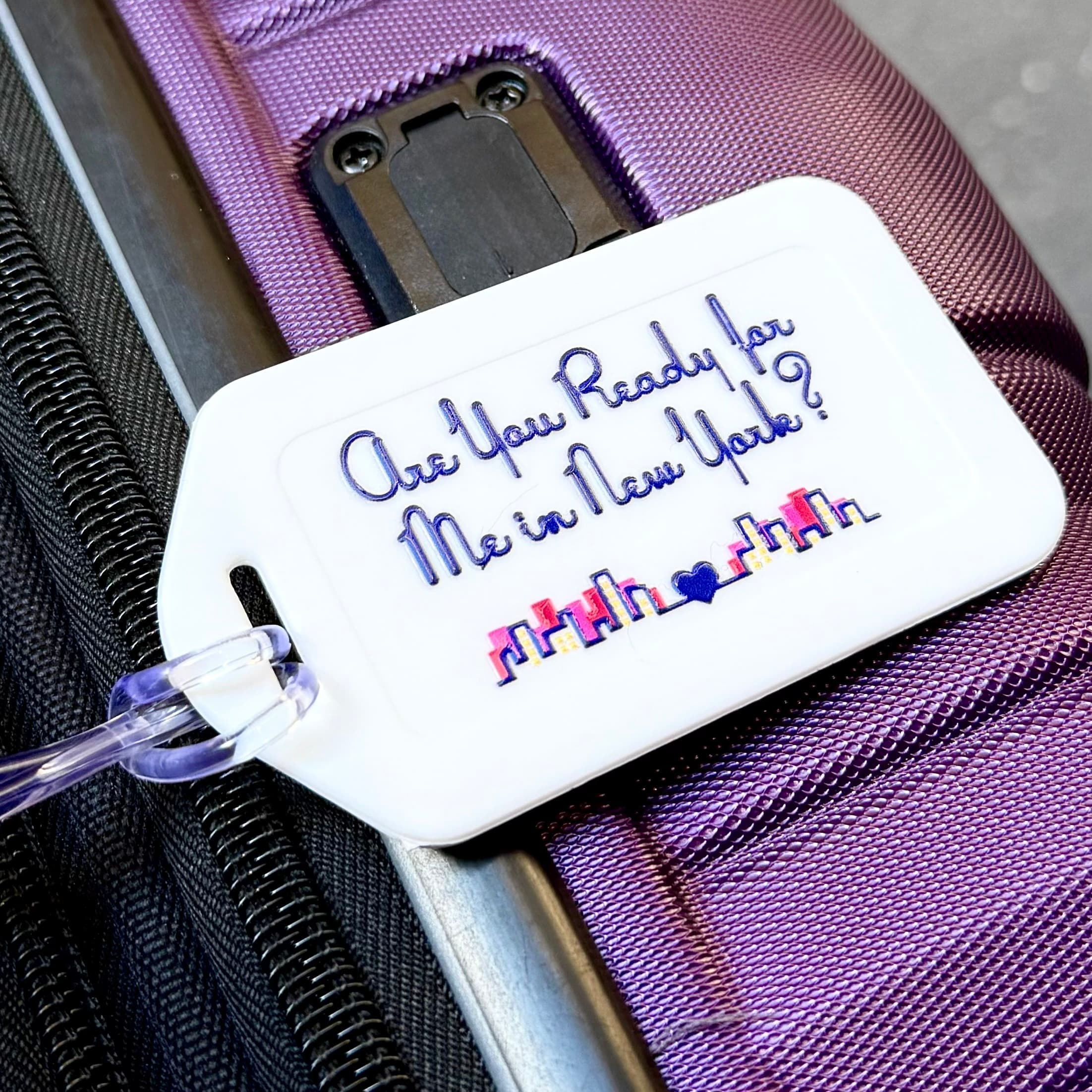 White luggage tag reading "Are You Ready for Me in New York?" with skyline, on purple suitcase.